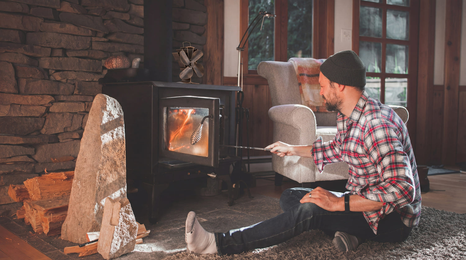 Man tending a wood fire in a cast-iron stove with a Breezy Stove heat-powered fan running on top, inside a cozy cabin living room with stone walls